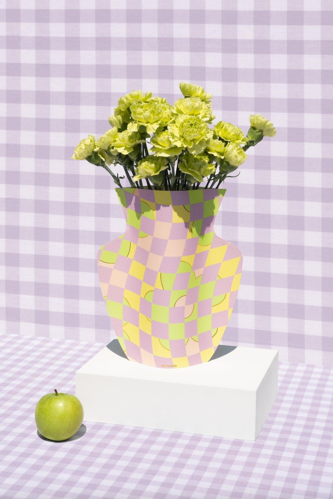 Yellow and pink checkered paper vase with yellow flowers on white pedestal, green apple beside it, purple gingham background