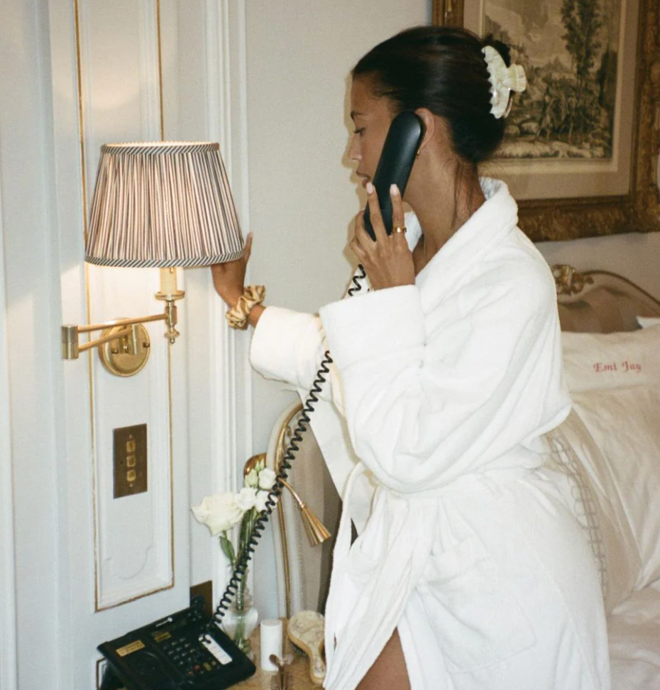 Woman in white bathrobe talking on corded telephone with flower in hair in elegant hotel room setting