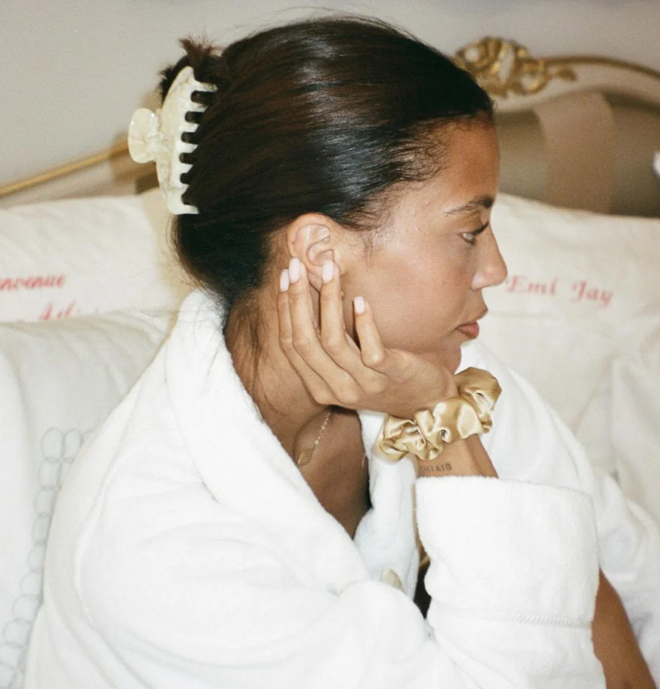 Woman in white robe with hair in bun secured by cream claw clip, wearing gold scrunchie bracelet while doing skincare routine