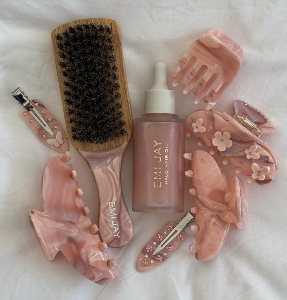 Pink hair care tools and accessories including brush, clips, claw clip, and Emi Jay hair oil bottle on white fabric