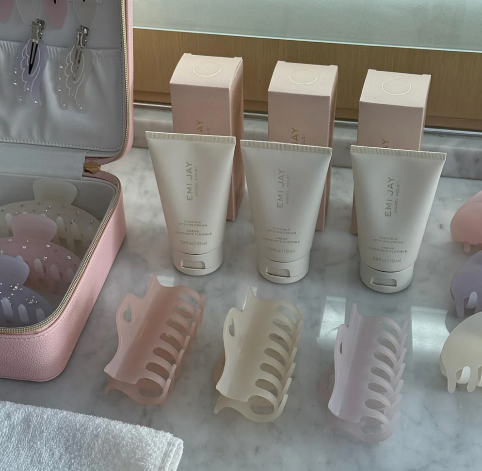 Three beige EMI JAY hair care tubes with matching boxes and claw clips displayed on marble surface with pink cosmetic bags