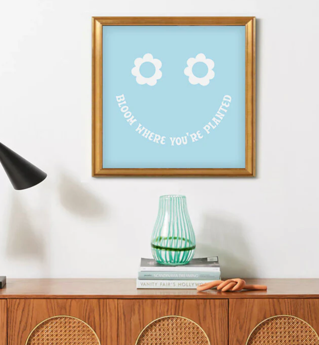 Framed blue wall art with white flower eyes and text "BLOOM WHERE YOU'RE PLANTED" forming a smile in a gold frame