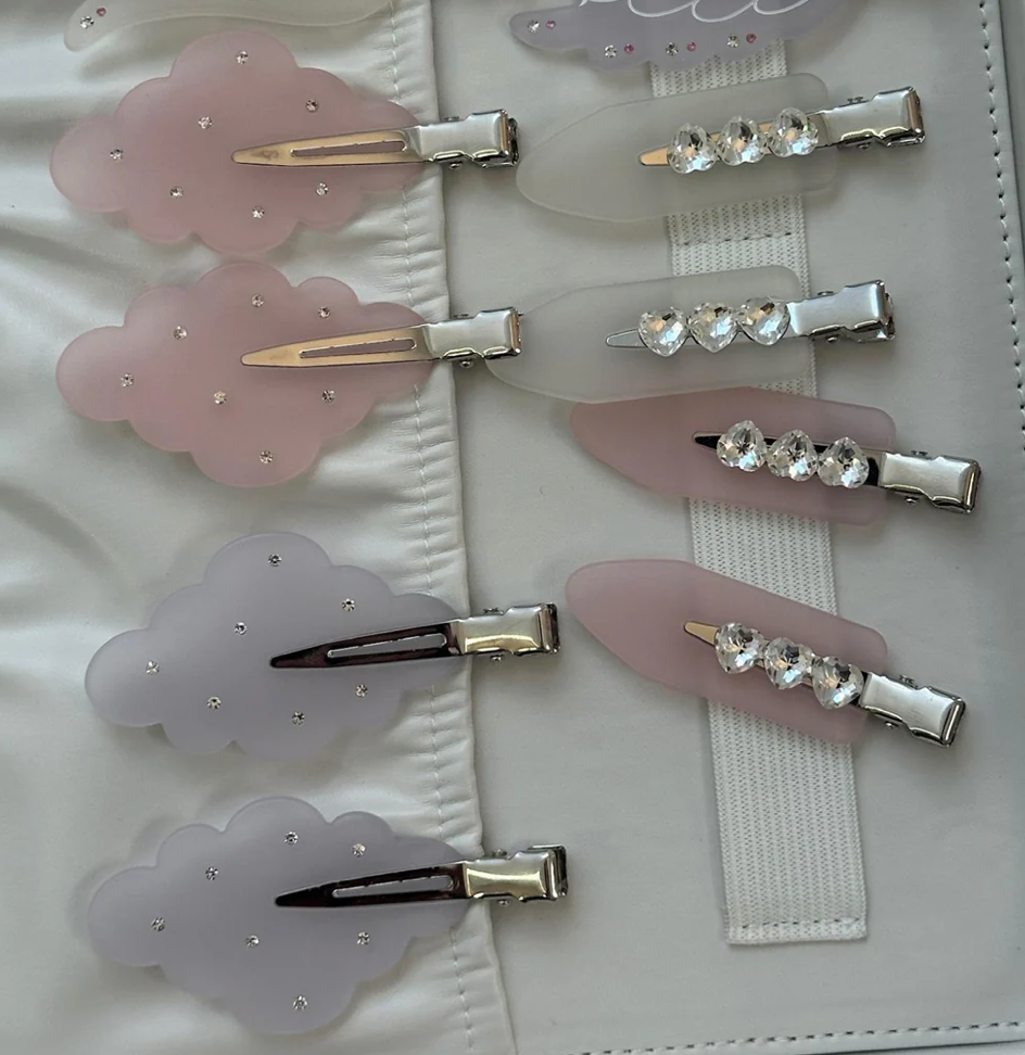 Pastel pink and lilac cloud-shaped hair clips with rhinestone details and silver metal clips, arranged on white fabric