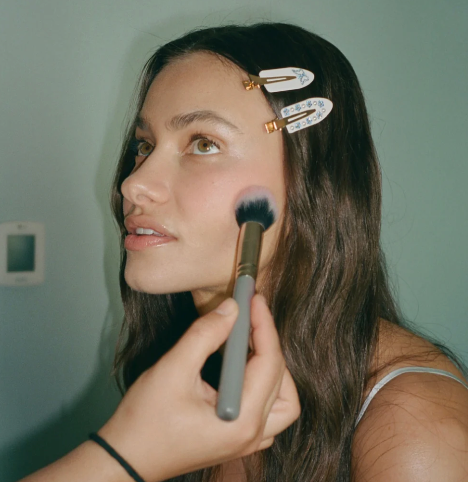 Woman applying makeup with brush, wearing white floral hair clips with gold accents in dark brown hair