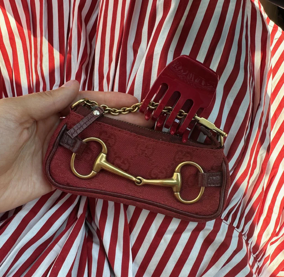 Red canvas shoulder bag with gold horsebit hardware and hair comb accessory held against red and white striped shirt