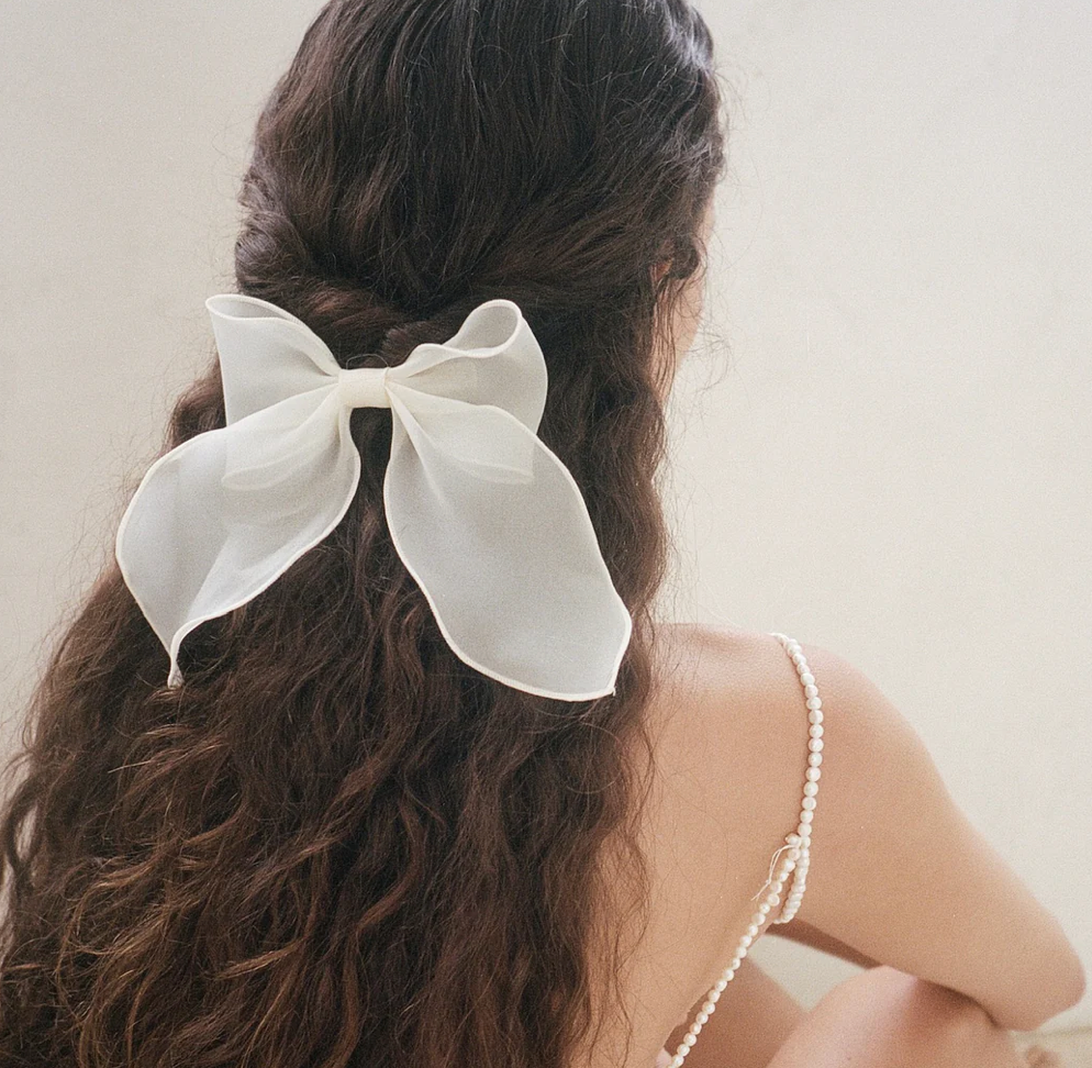 Large white fabric bow hair clip with flowing ribbons secured in brunette wavy hair, half-up hairstyle