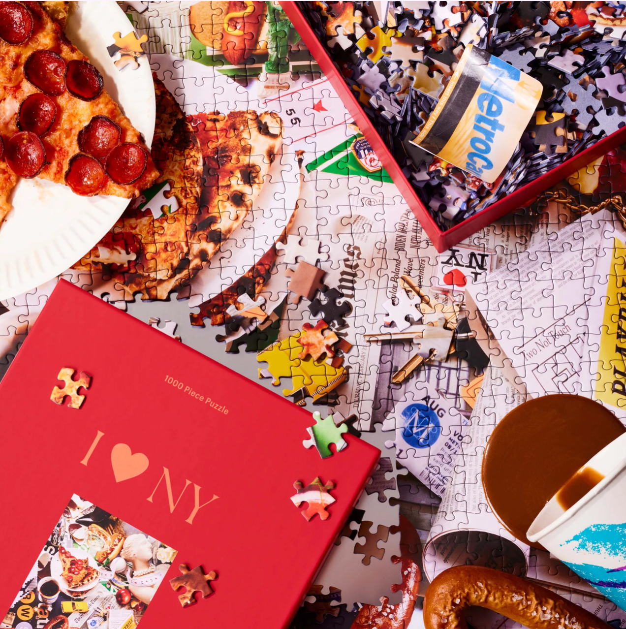 New York themed jigsaw puzzles with red I Love NY box, scattered puzzle pieces, and pepperoni pizza slice on white background