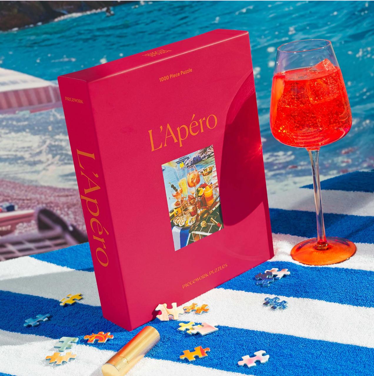 Red puzzle box labeled L'Apéro by a pool with scattered puzzle pieces, wine glass with orange drink, and striped towel