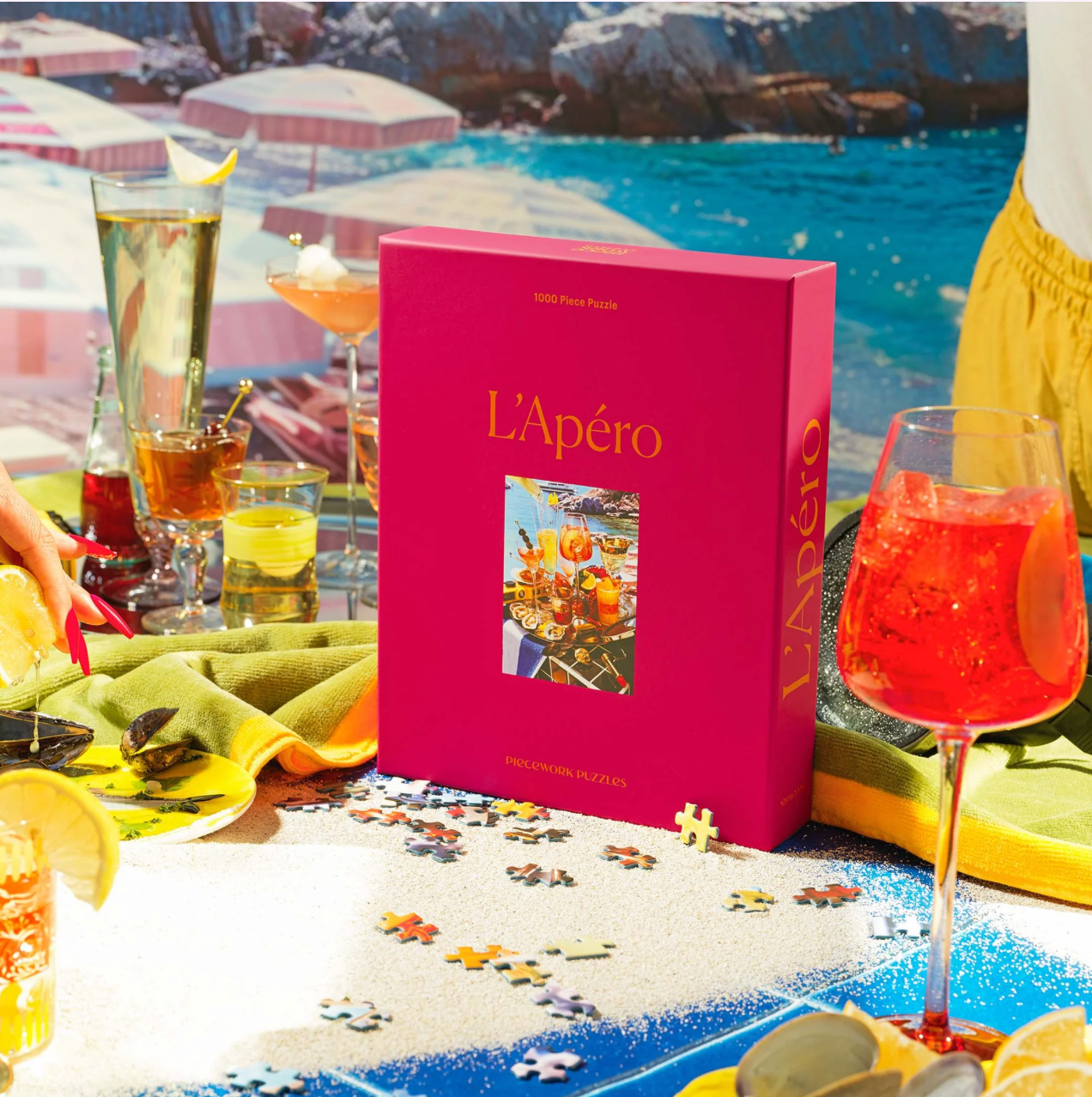 Pink puzzle box labeled L'Apéro featuring cocktails scene, displayed on beach setting with drinks and scattered puzzle pieces