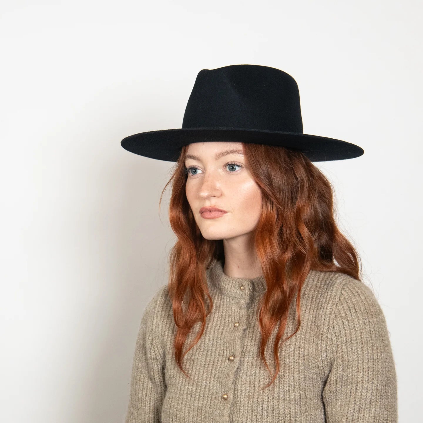 Black wide-brim fedora hat worn by woman with long wavy auburn hair and beige knit sweater against white background