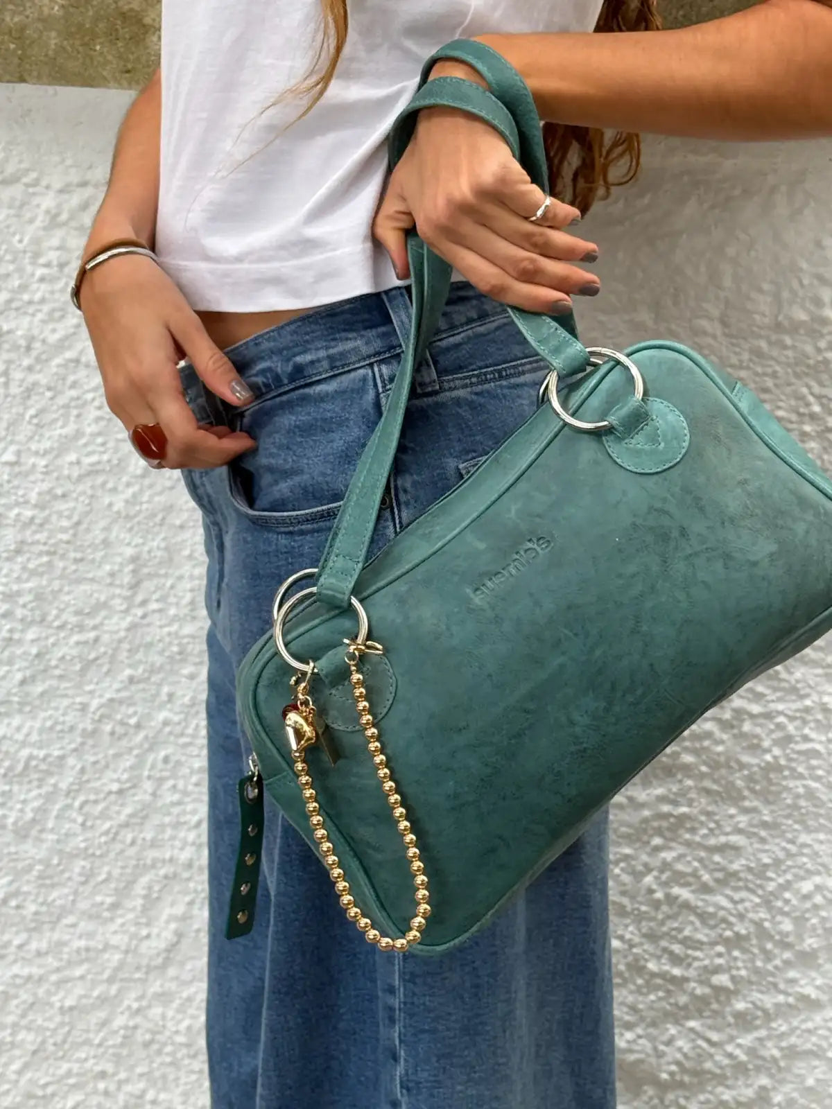 Teal green suede handbag with silver ring hardware and gold beaded chain detail, carried by woman in white tee and jeans