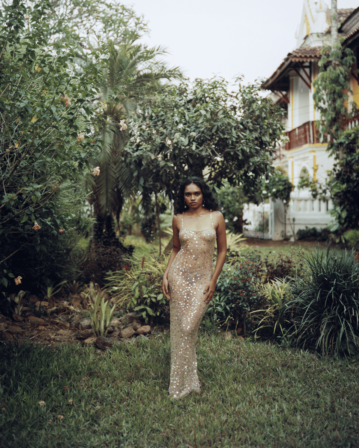 Woman in gold sequined floor-length gown standing in lush garden with tropical plants and colonial-style house in background