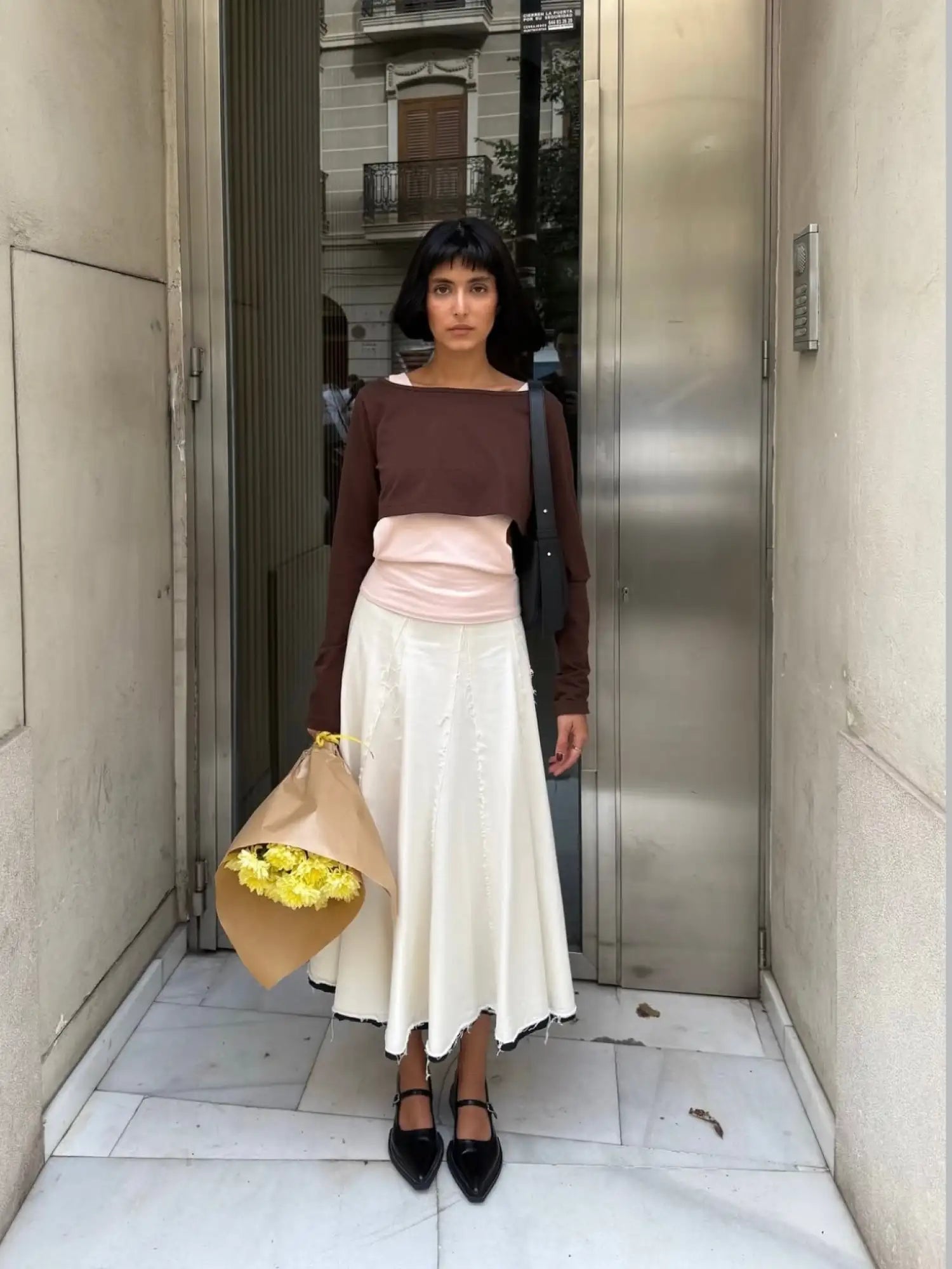 Woman in brown long-sleeve top and white midi skirt holding yellow flowers in tan paper wrapping, wearing black Mary Jane shoes