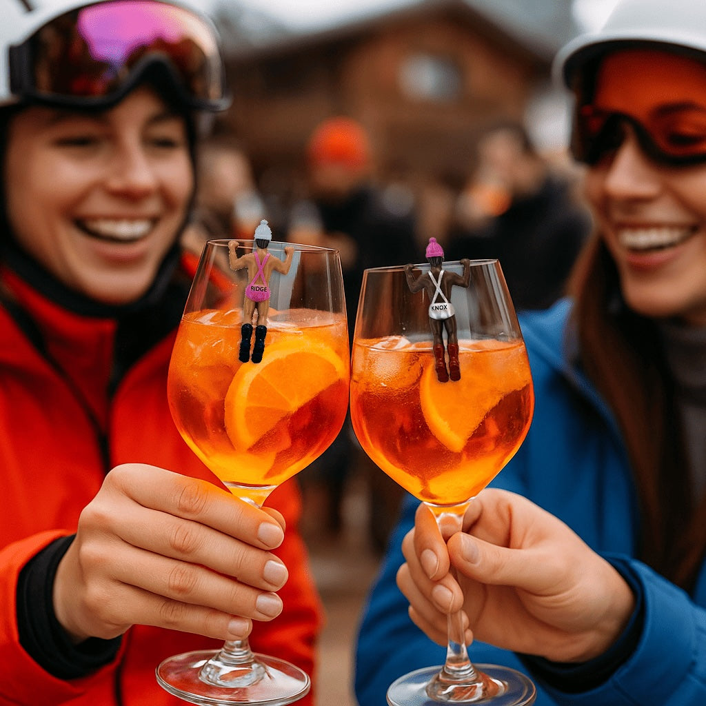 Two people in ski gear toasting orange cocktails with miniature skier figurines perched on glass rims at winter resort
