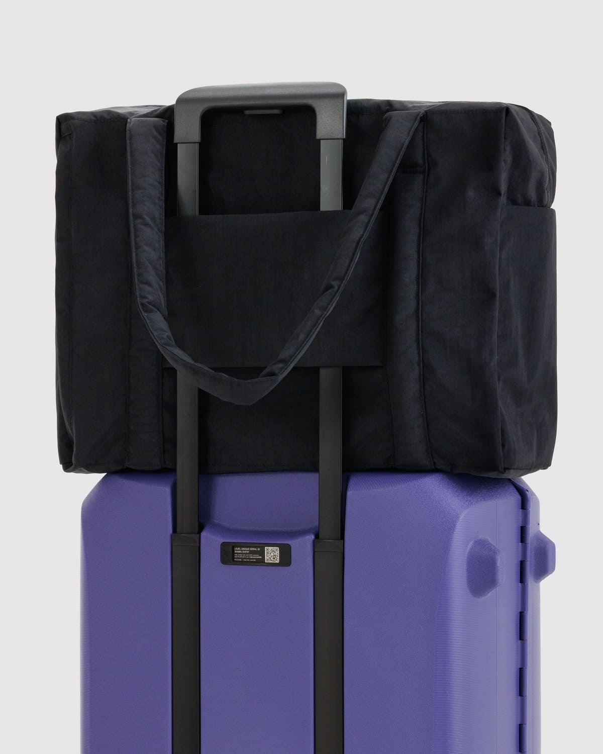 Black duffel bag with shoulder straps attached to purple hardshell suitcase via luggage sleeve and telescopic handle