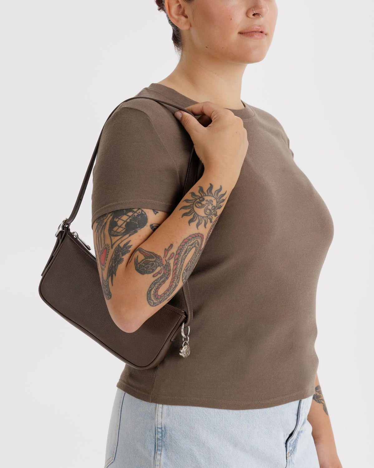 Brown leather shoulder bag with silver hardware worn by tattooed model in olive tee and light jeans