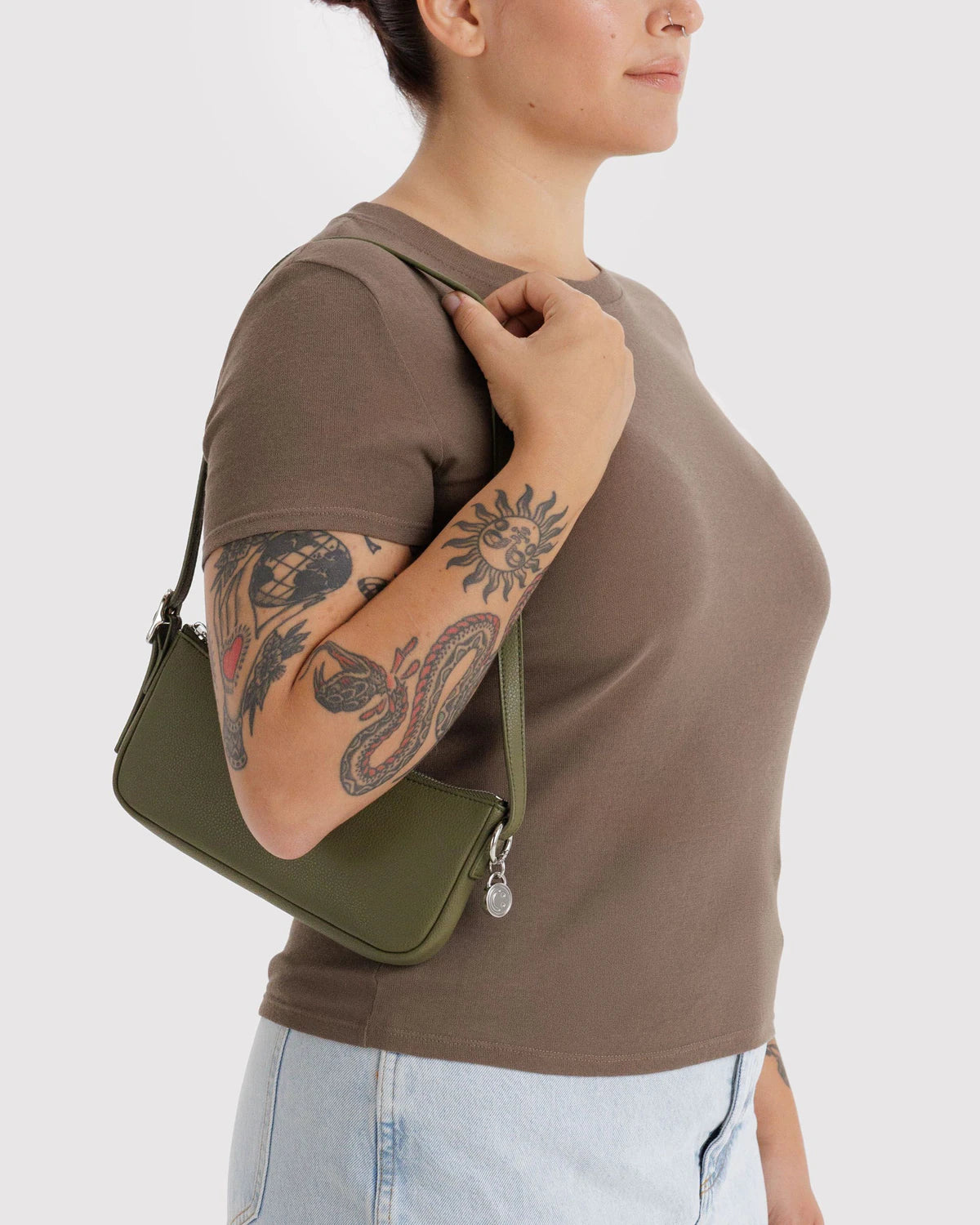 Olive green mini shoulder bag with adjustable strap worn by tattooed model in brown long-sleeve top