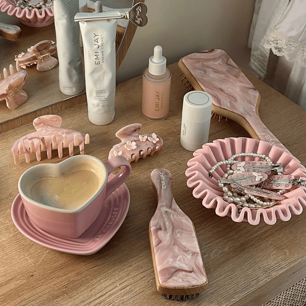 Pink marble beauty and hair care flatlay with EMI JAY products, coffee cup, hair clips, brush, and accessories on wood table
