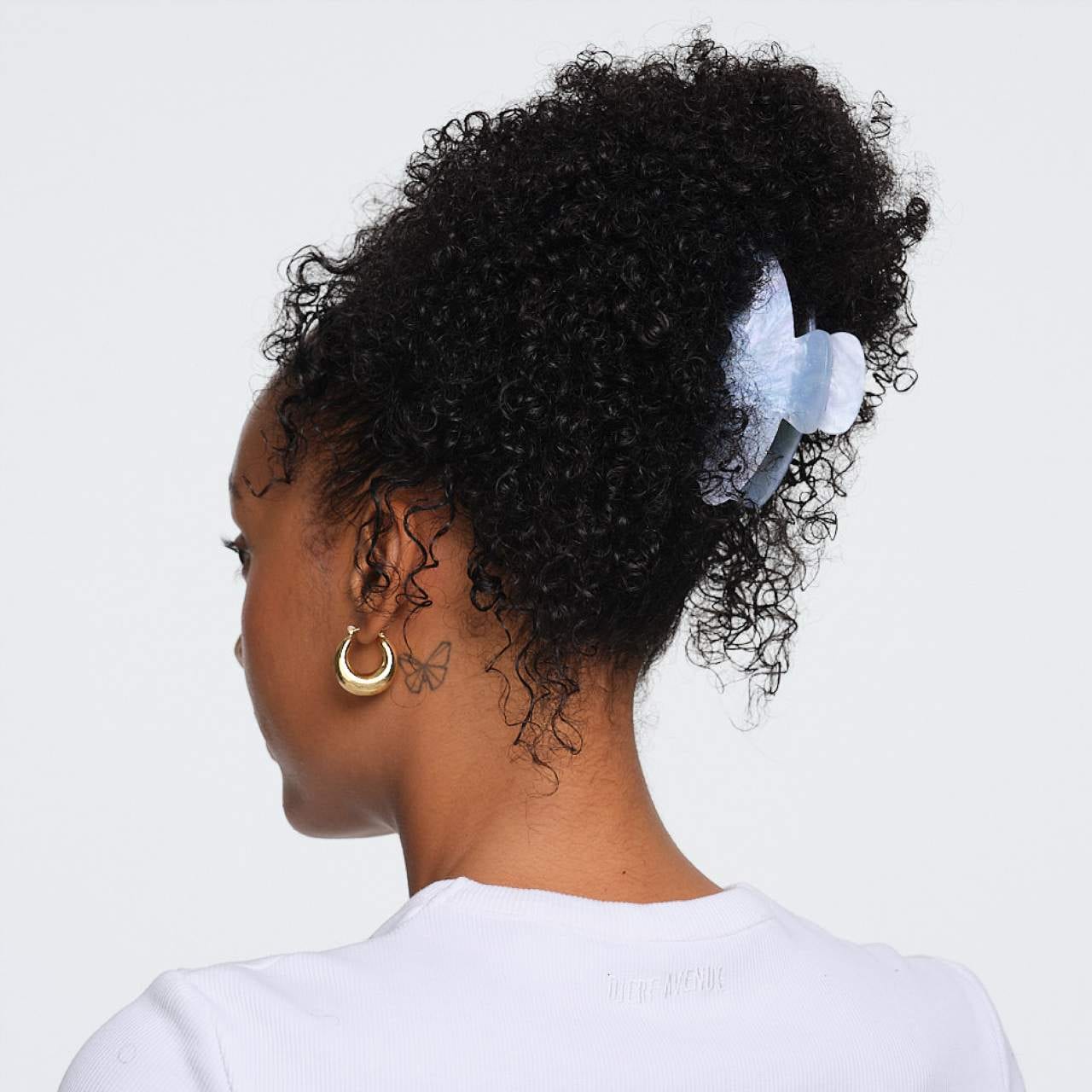 Large gold hoop earring on model with curly black hair in white top, shown from side/back view