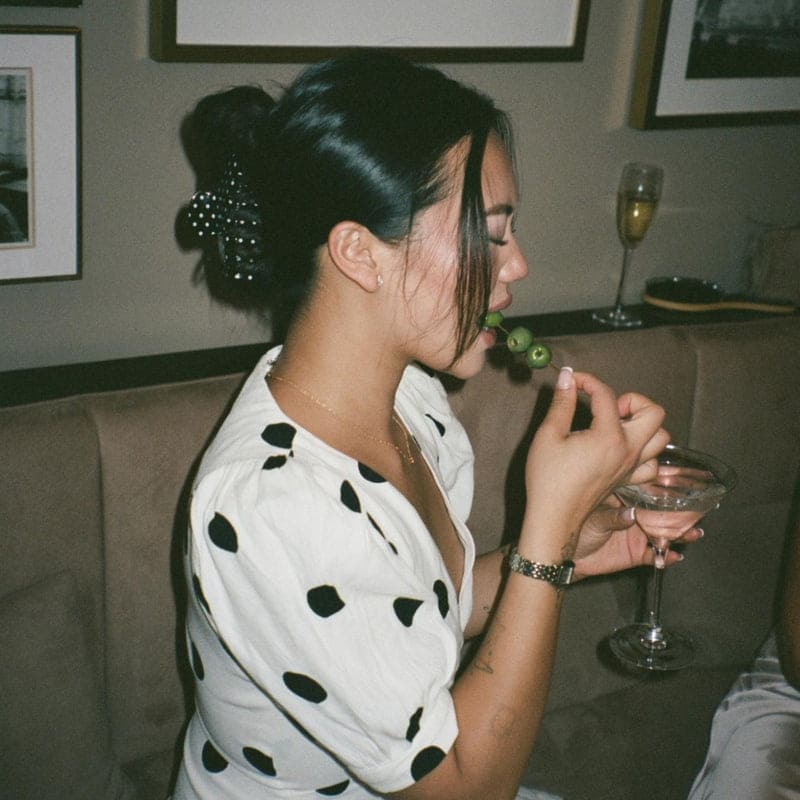 Woman wearing white button-up shirt with black polka dots, holding cocktail glass with decorative hair clip