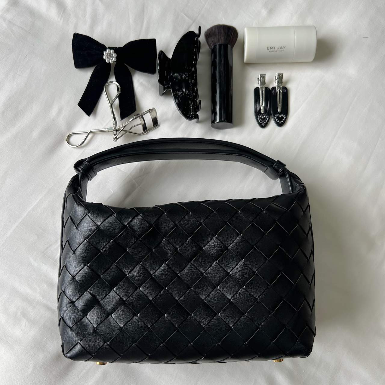 Black woven leather shoulder bag displayed with makeup accessories including brushes, bow clip, eyelash curler and nail clippers on white fabric