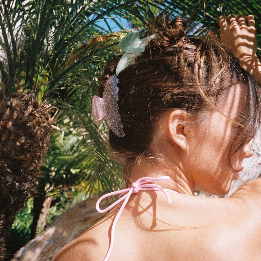 Glittery purple butterfly hair clip in brunette hair with palm trees in background, pink bikini top visible