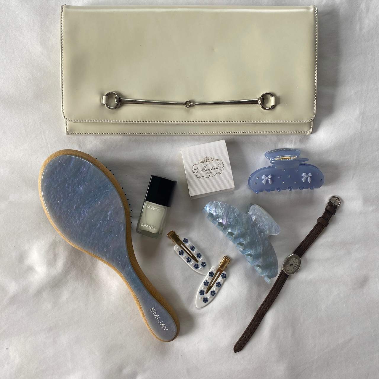 Cream leather clutch with silver horsebit detail displayed with blue hair accessories, nail polish, hairbrush and watch