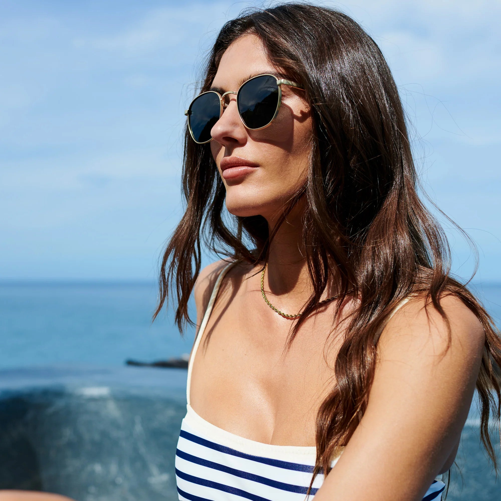 Woman wearing sunglasses and a striped swimsuit with a blue sky and ocean background