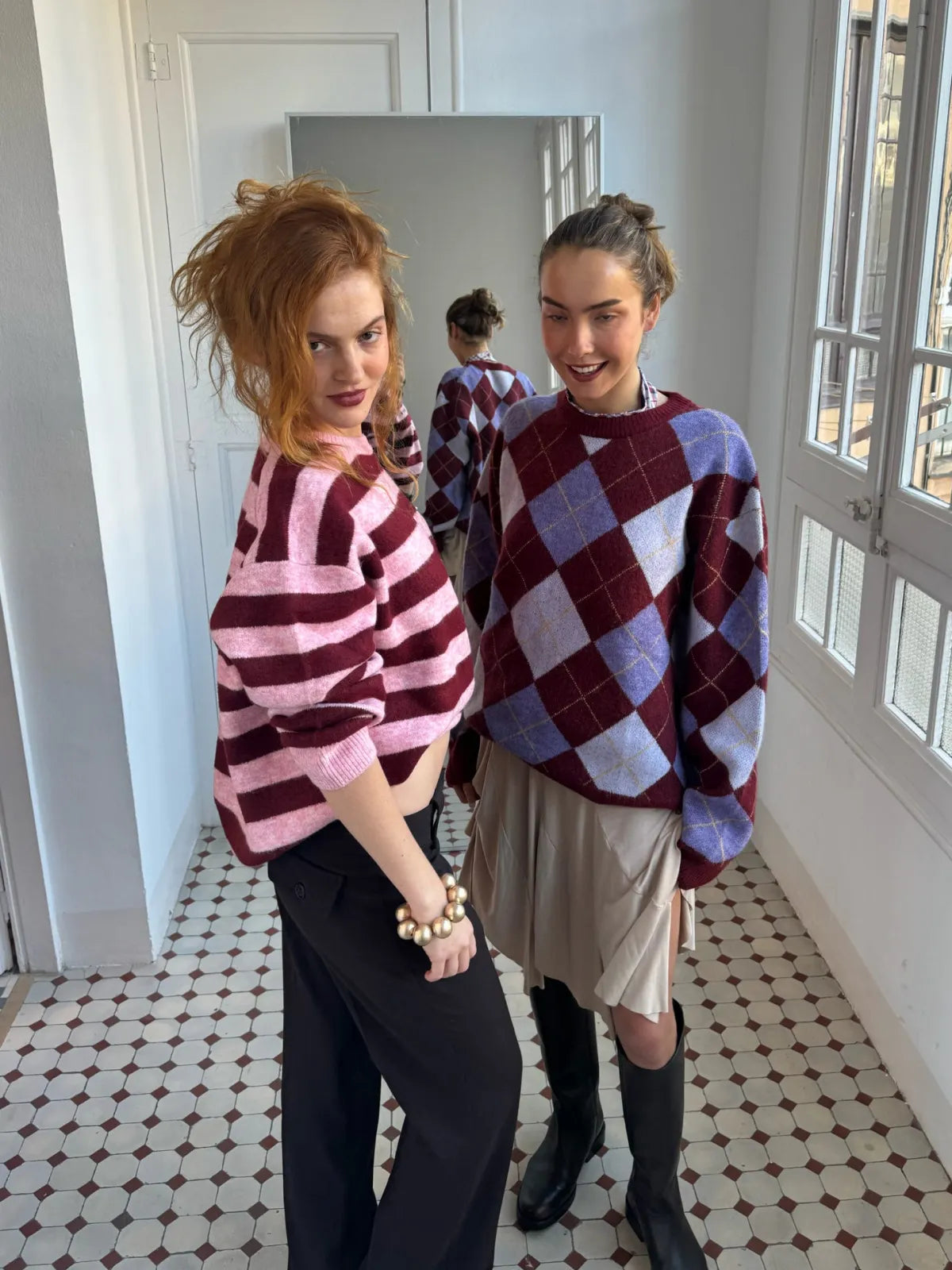Two women wearing coordinating burgundy and pink patterned sweaters - one striped, one argyle - styled with pants and boots