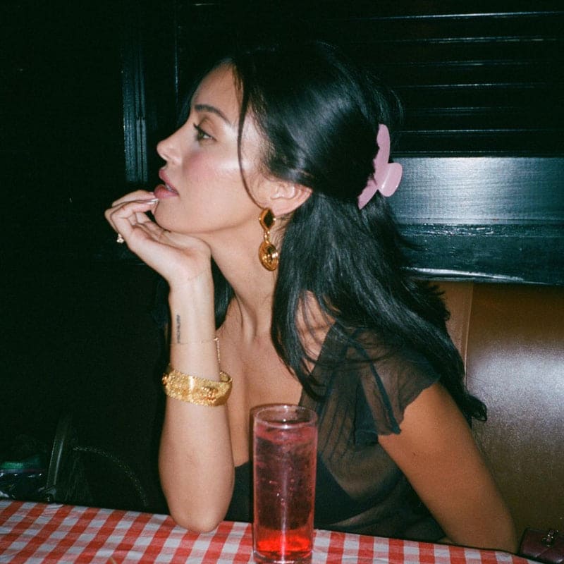 Woman with dark hair wearing gold statement earrings and bracelet, pink hair clip, seated at table with red drink in diner booth