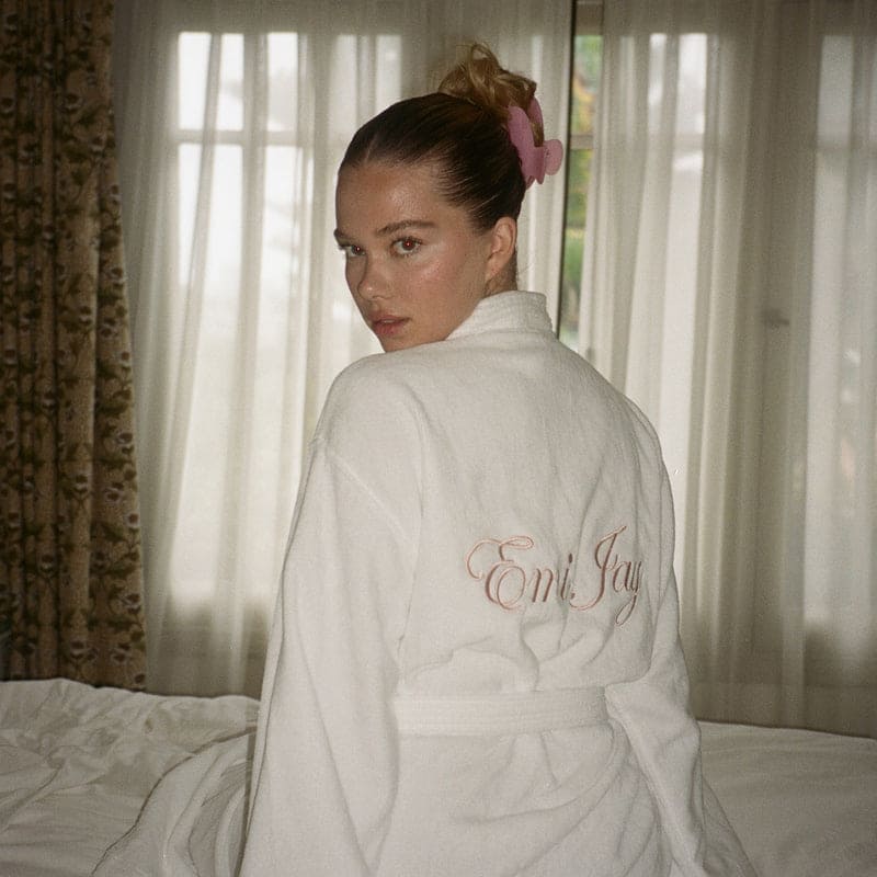 White cotton robe with 'Emi Jay' embroidered in pink script on back, worn by woman with hair bow