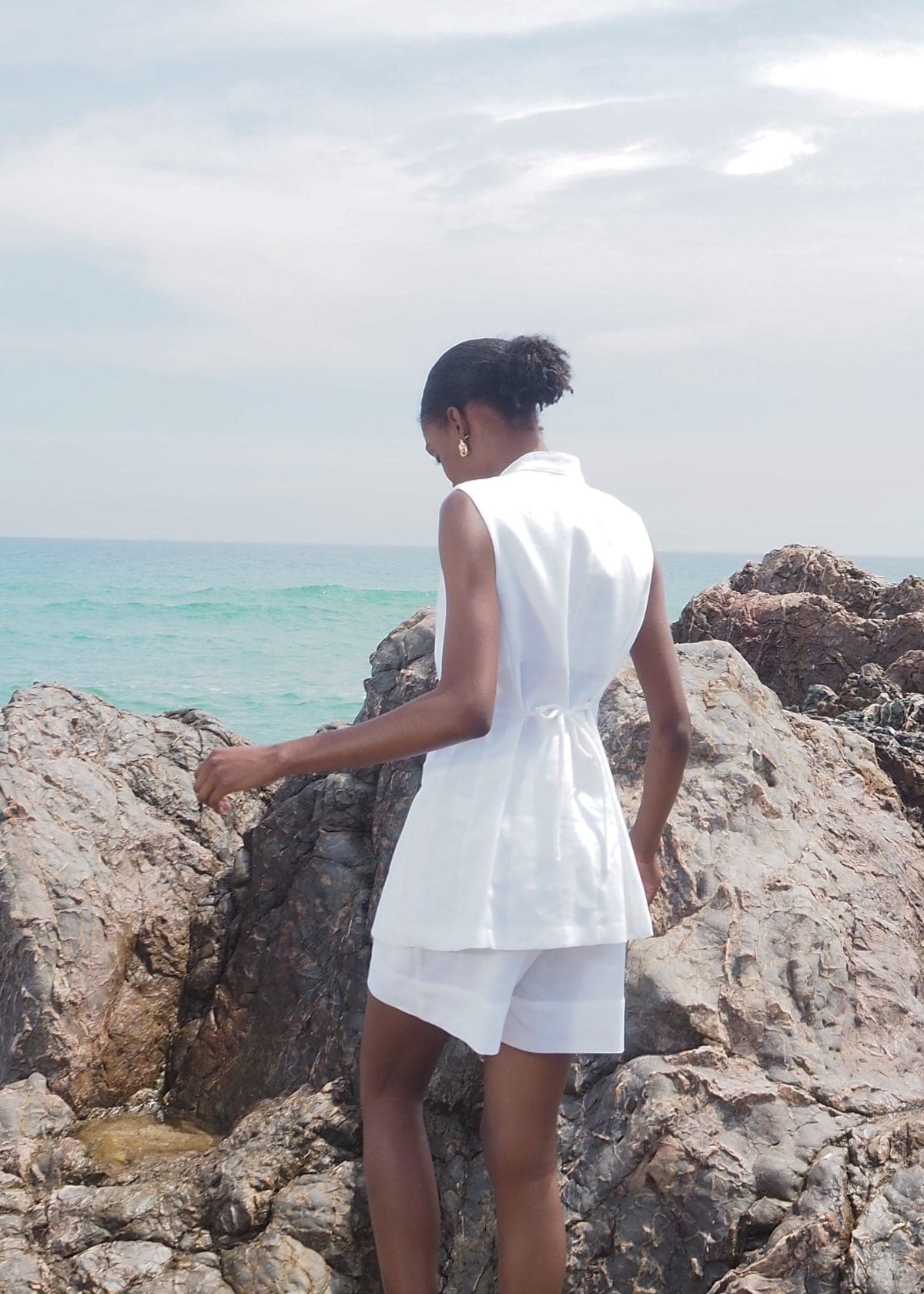 Woman in white sleeveless belted shirt dress standing on rocky coastline overlooking turquoise ocean water