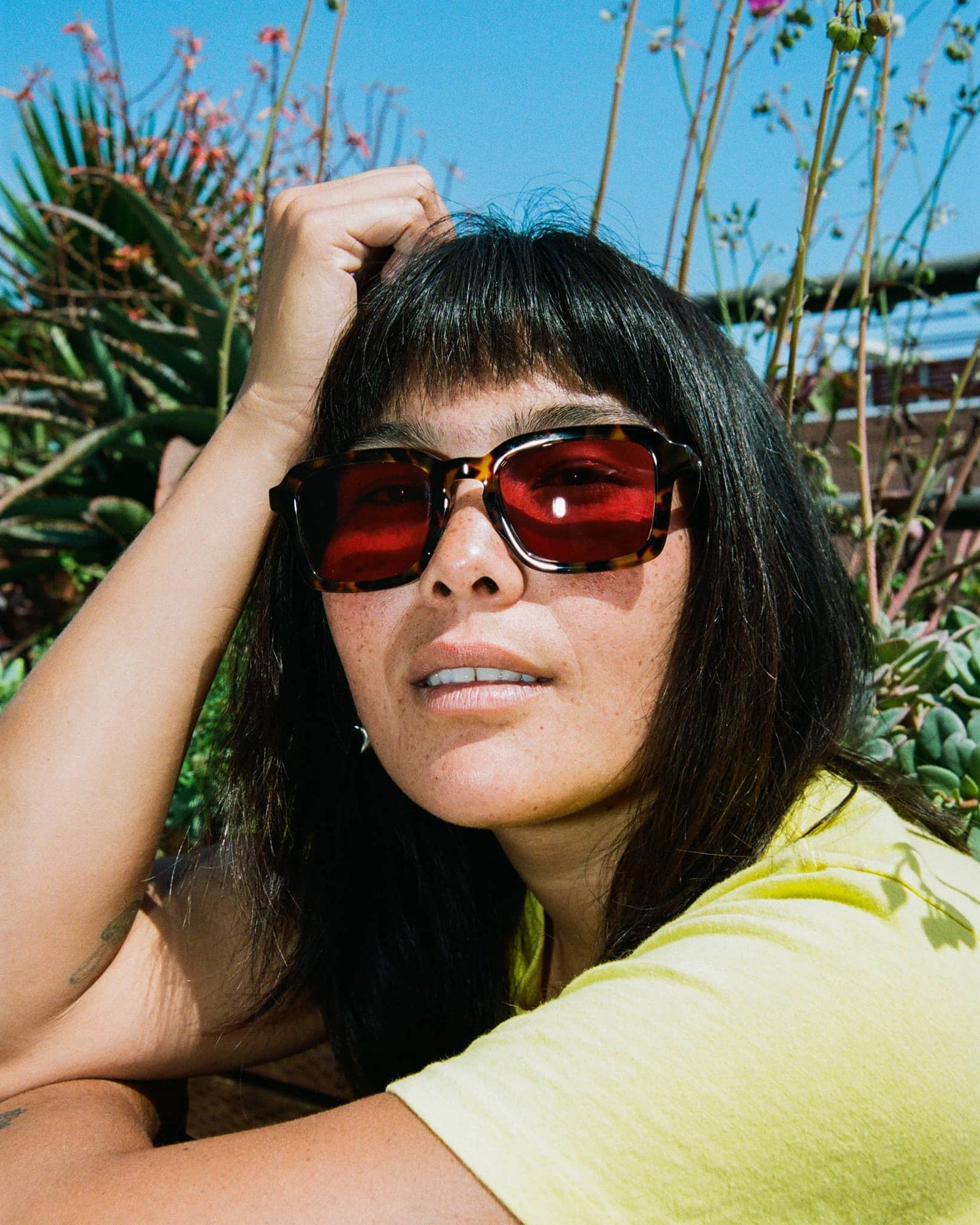 Woman wearing tortoiseshell sunglasses with red-tinted lenses and yellow-green shirt outdoors among garden plants