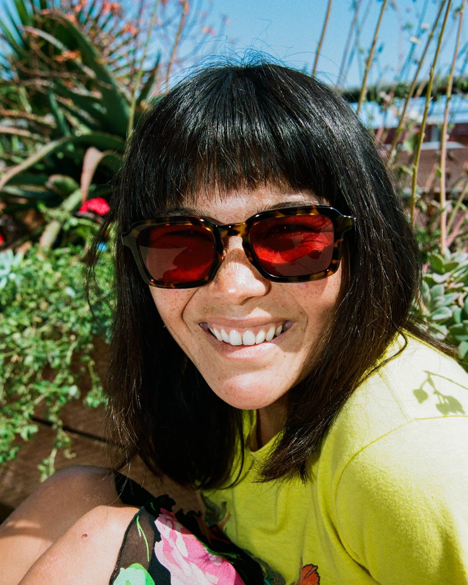 Woman wearing tortoiseshell sunglasses with red-tinted lenses and black hair with bangs, smiling outdoors in bright sunlight