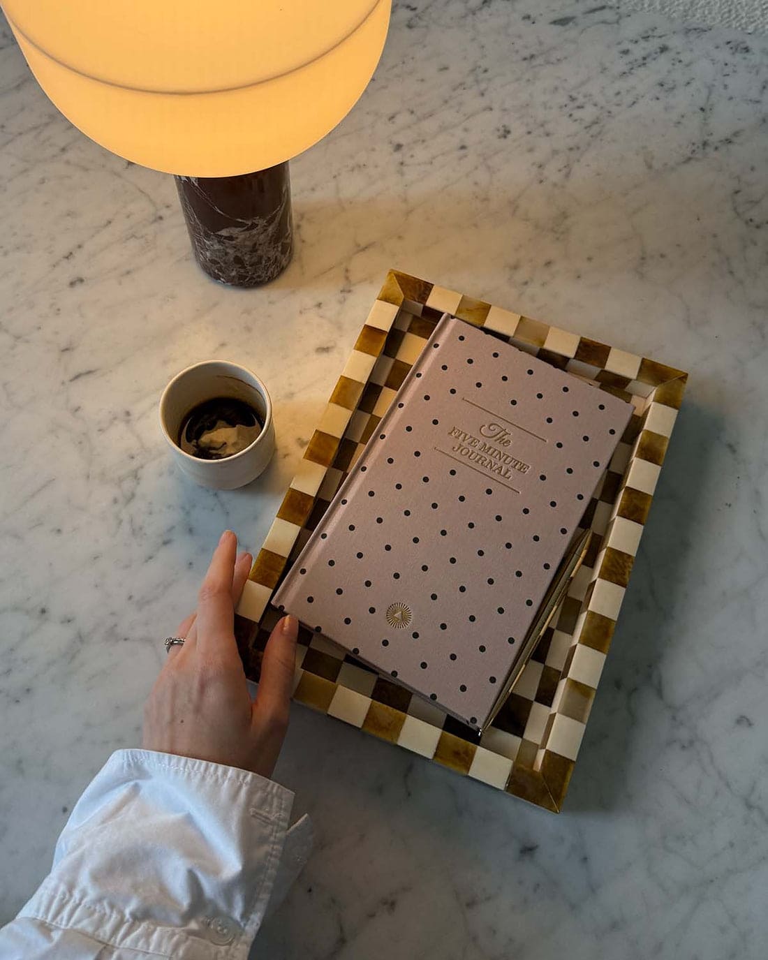Checkered bone inlay tray in gold and cream tones holding a polka dot journal, displayed on marble surface with coffee cup