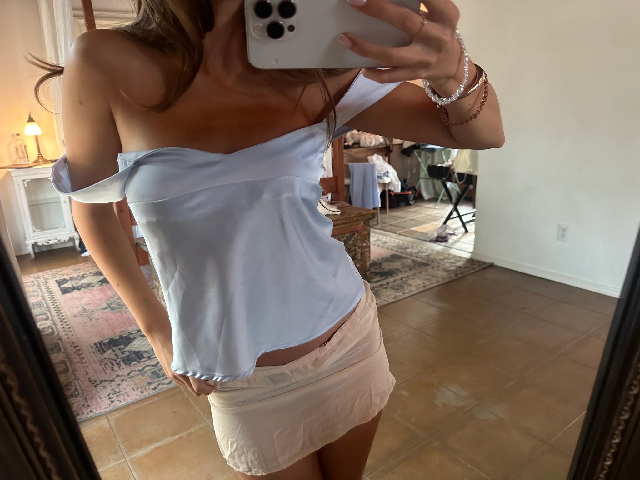 Person taking a mirror selfie wearing a white top and beige skirt in a room.
