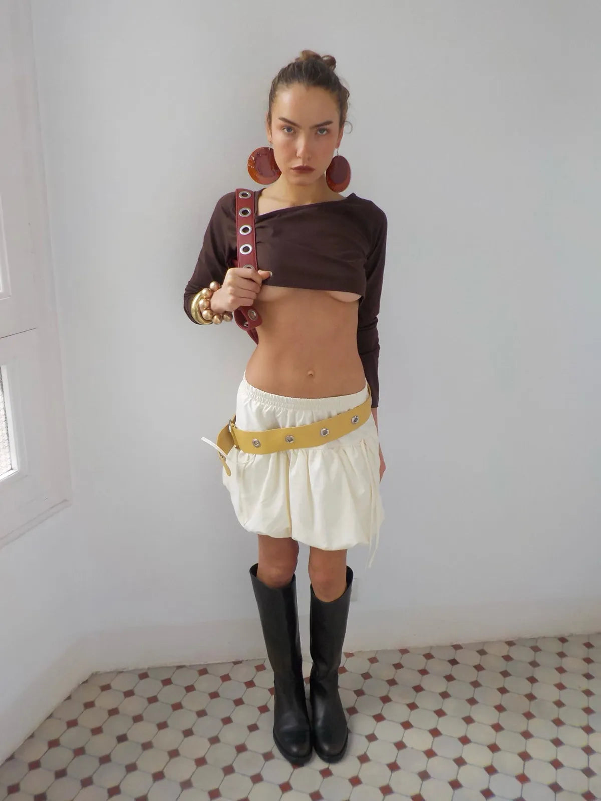 Woman modeling avant-garde outfit with brown crop top, cream shorts with yellow belt, oversized wooden earrings, and black boots