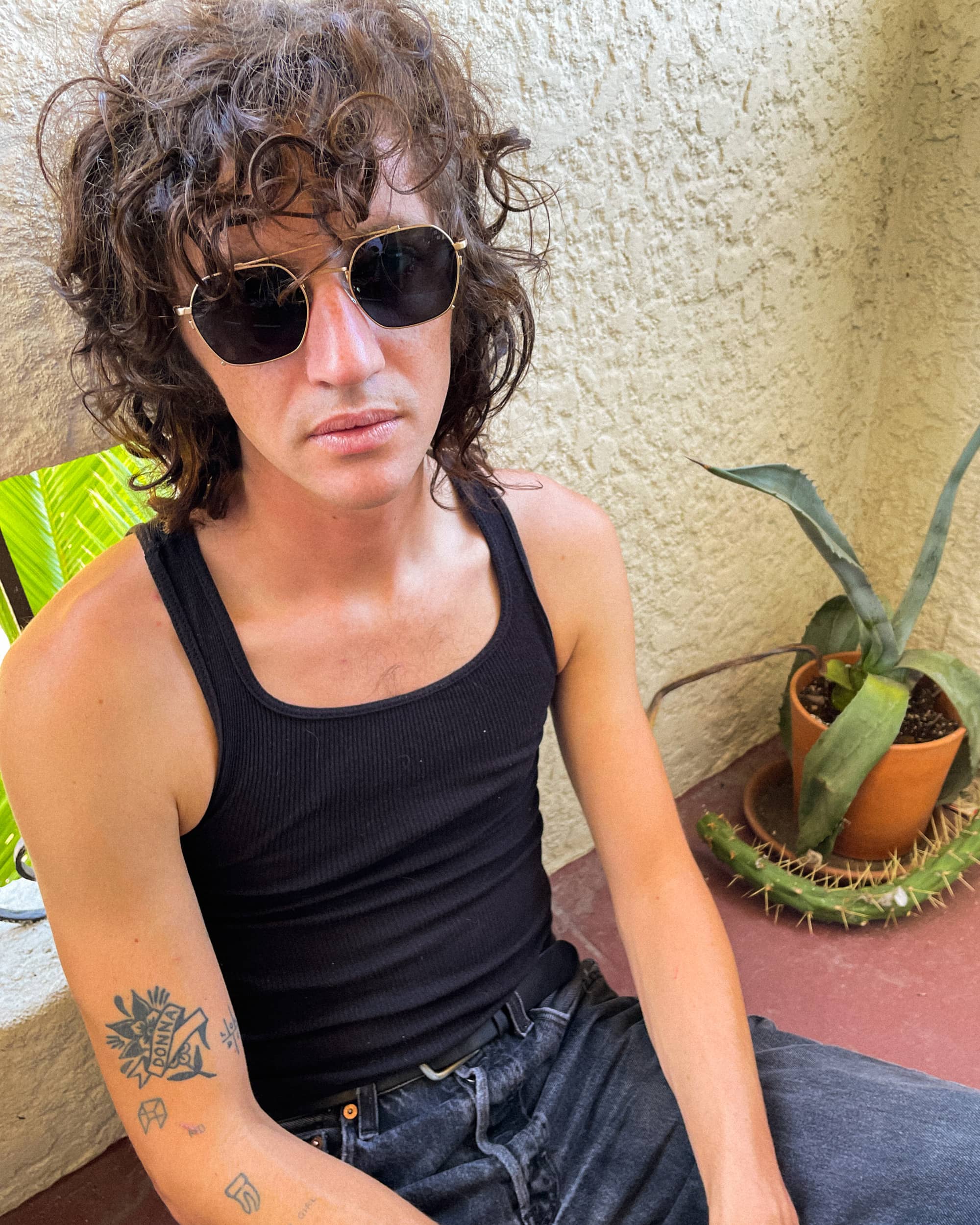 Person with curly hair wearing sunglasses and a black tank top, sitting outdoors.