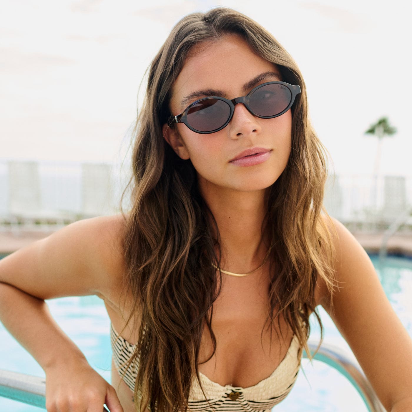 Woman wearing black oval sunglasses by a pool, featuring dark tinted lenses and sleek retro-style frames