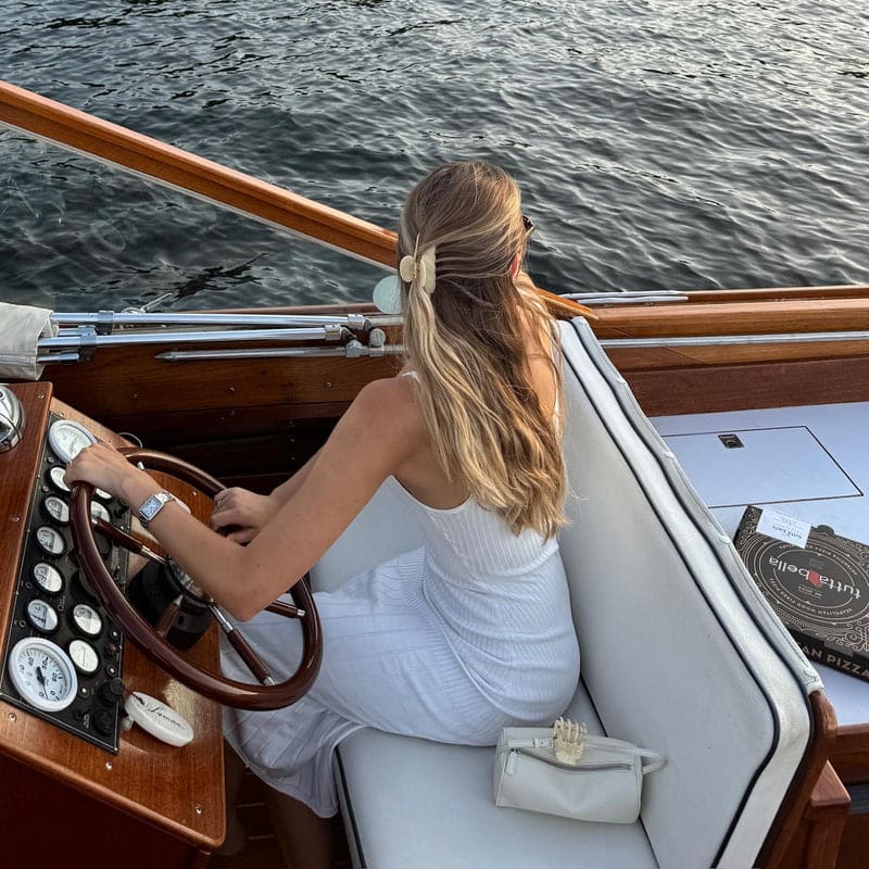 Woman in white dress steering wooden boat on water, view from behind showing long blonde hair with bow clip