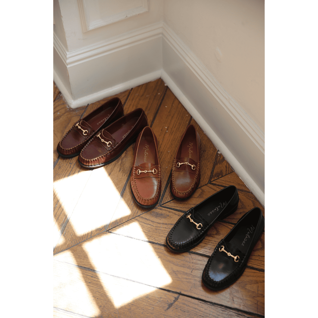 Three pairs of leather loafers with gold horsebit hardware in brown, tan, and black colors arranged on wooden floor