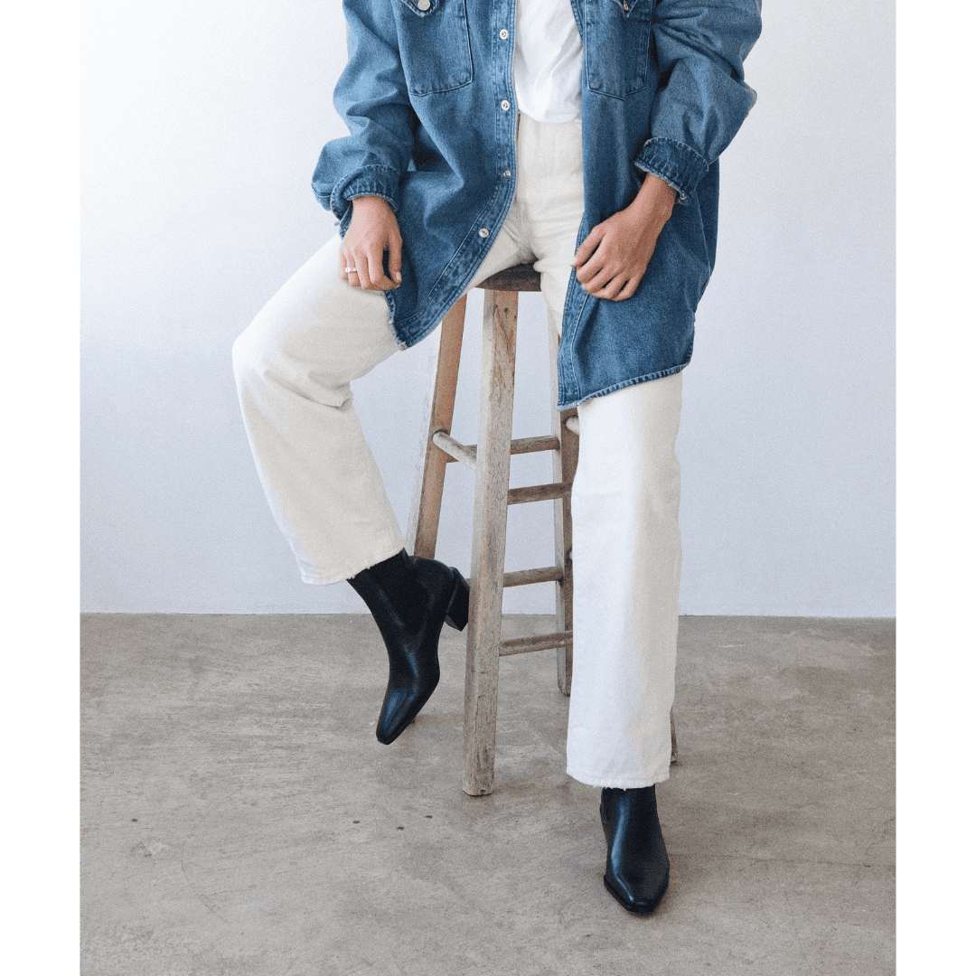Person wearing cream wide-leg pants with denim shirt and black ankle boots, sitting on wooden stool