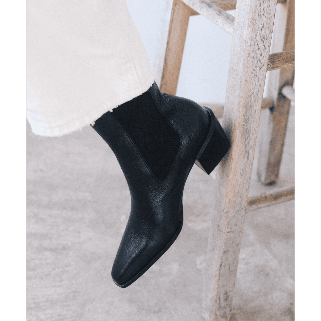 Black leather ankle boots with block heel on wooden chair, worn with white pants