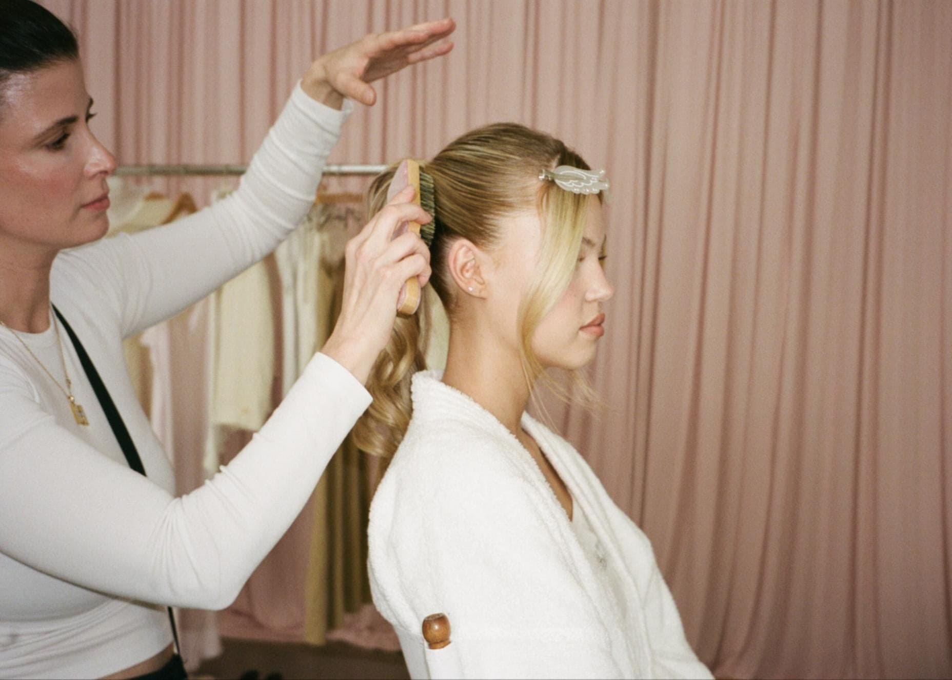 Hairstylist styling blonde woman's hair with decorative pearl clip in white robe against pink curtain backdrop