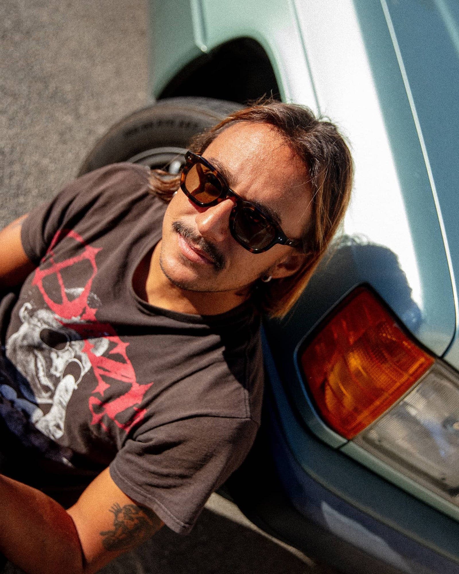Man wearing tortoiseshell sunglasses and graphic t-shirt leaning against vintage car with teal and blue body panels