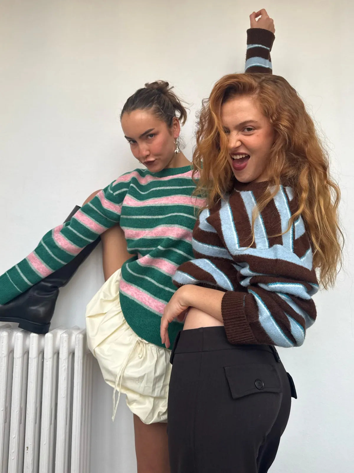 Two women wearing colorful striped sweaters - one in green and pink stripes, one in brown and blue stripes - posing playfully