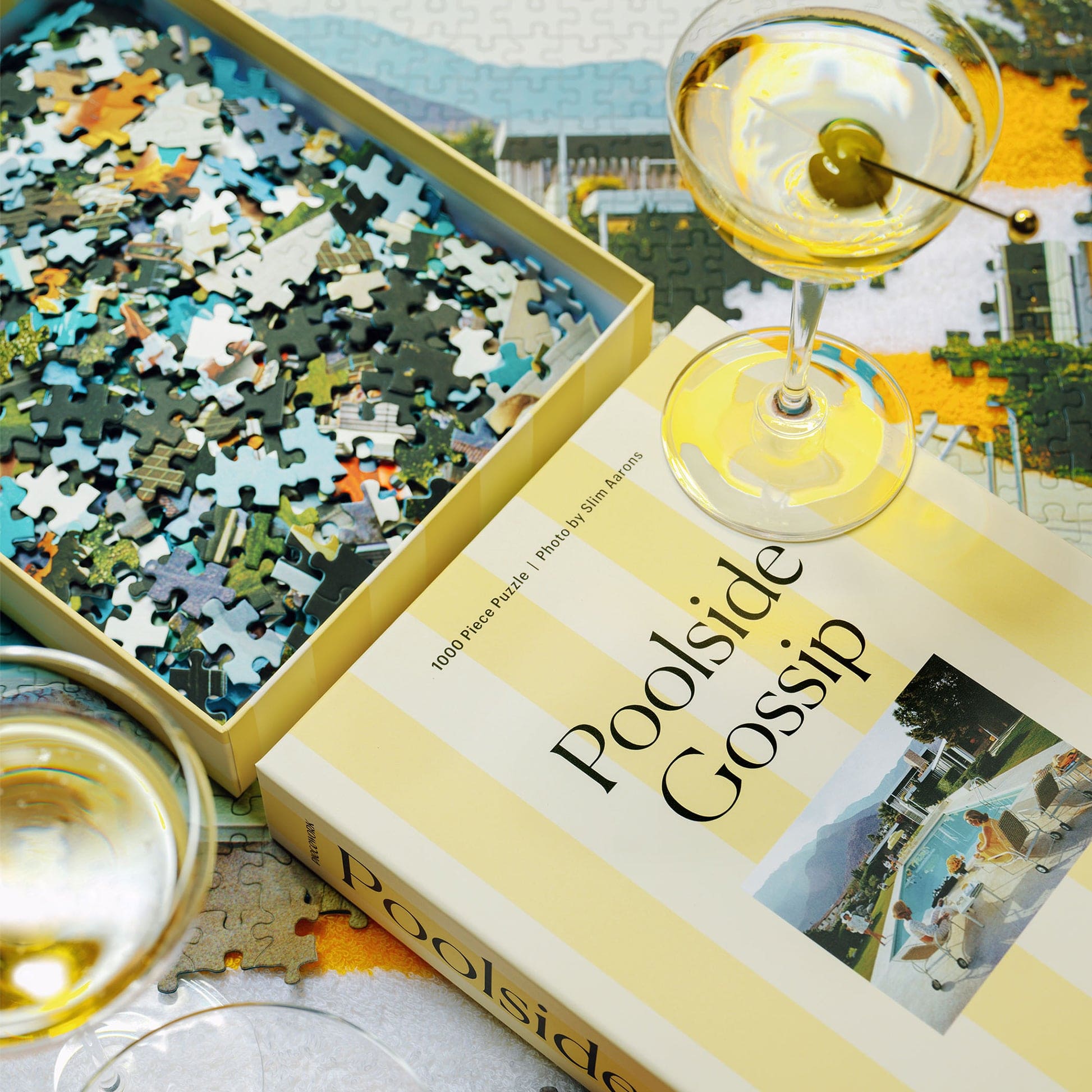 Poolside Gossip 1000-piece jigsaw puzzle in yellow box with scattered pieces and martini glasses on white surface