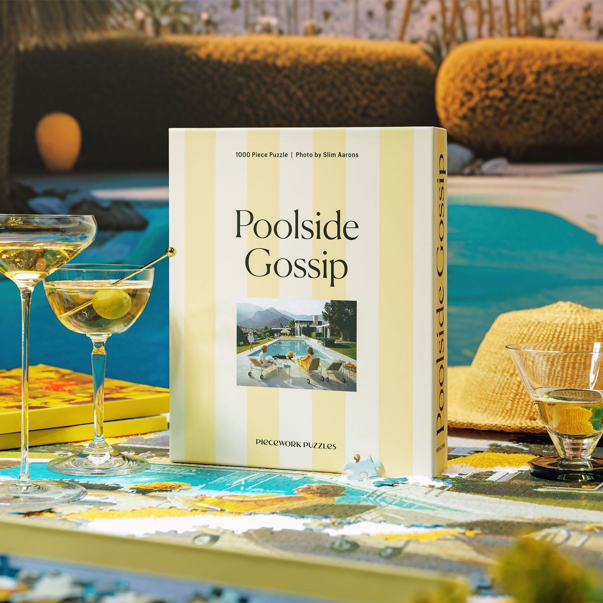 Poolside Gossip 1000-piece jigsaw puzzle box with yellow striped design, displayed on turquoise poolside table with cocktails