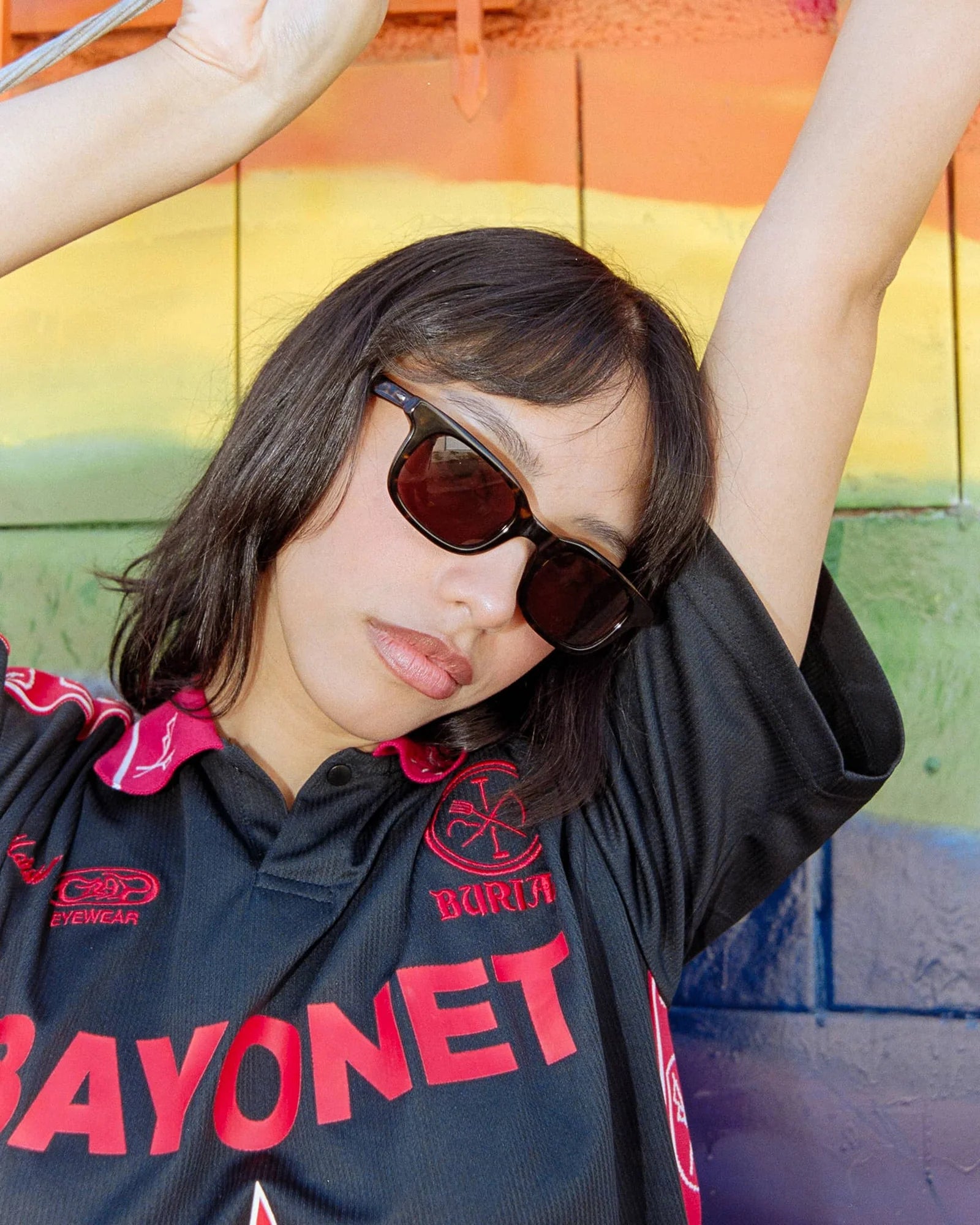 Person wearing sunglasses and a black shirt with red text against a colorful wall.