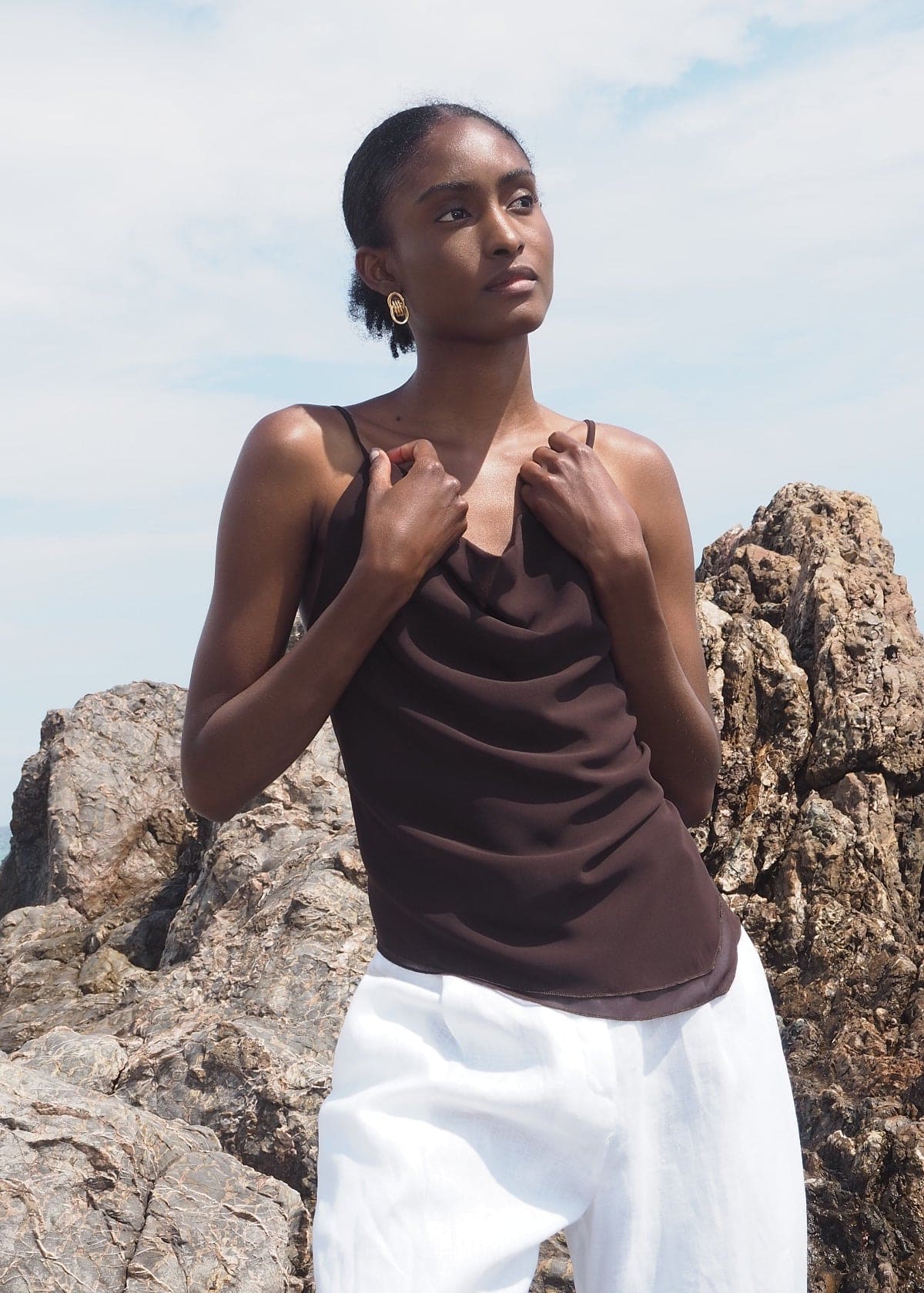 Woman wearing dark brown satin camisole with thin straps and white pants, standing outdoors against rocky backdrop